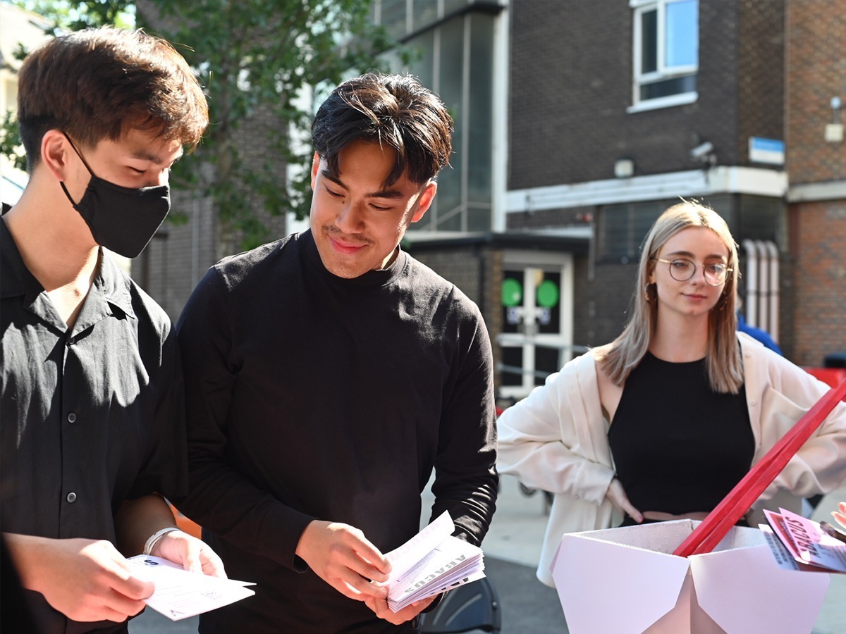 Two people are looking at different flyers. Another person is standing in the background looking at them.