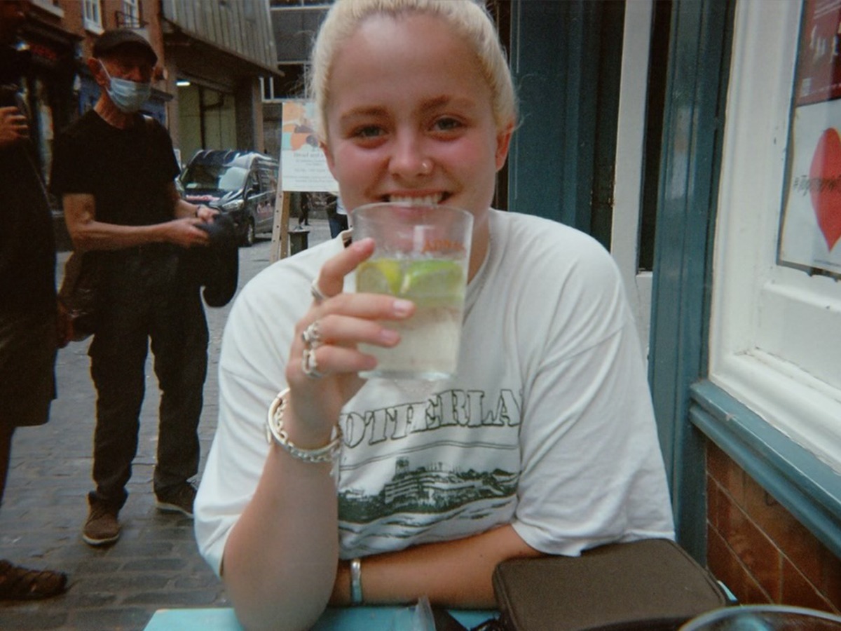 Reid is sitting down, holding a glass up to her mouth. She is wearing rings, has bleached hair and is smiling. 