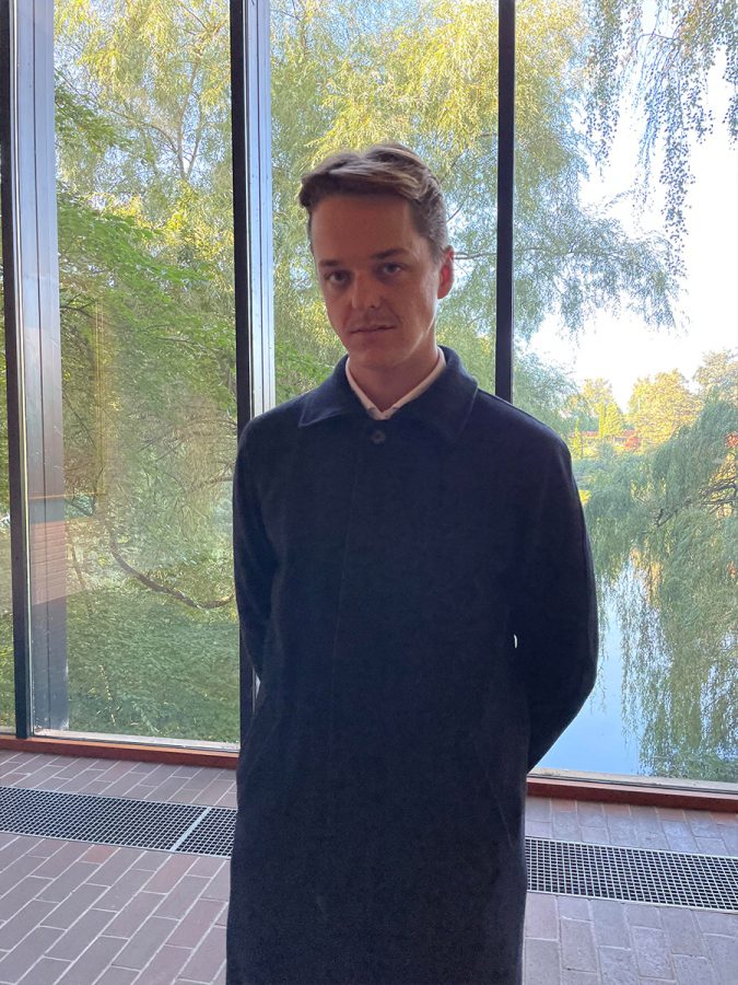 Frantzen is standing is front of a window. He is wearing a long coat and is staring into the camera. Outside, there is a lake surrounded by trees.