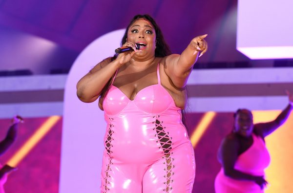 Lizzo wearing a pink latex jumpsuit on stage pointing at the crowd.