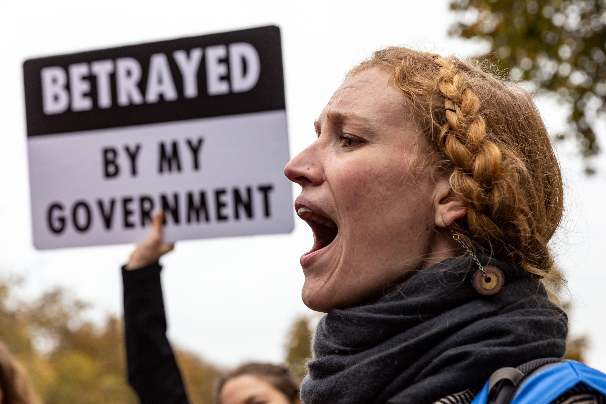 A person protesting with a sign reading "betrayed by the government" in the background