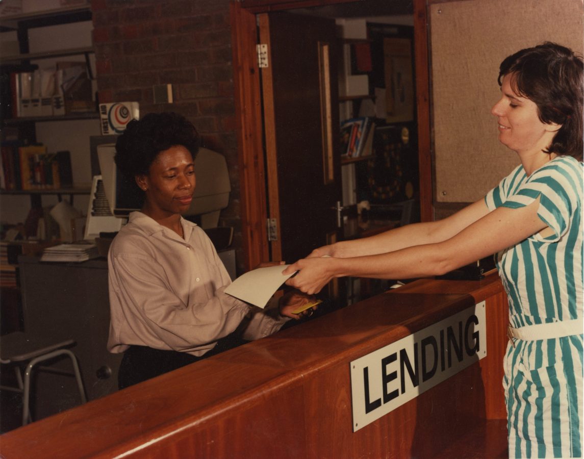 Woman sat behind a library lending desk, passing a book to a student. 