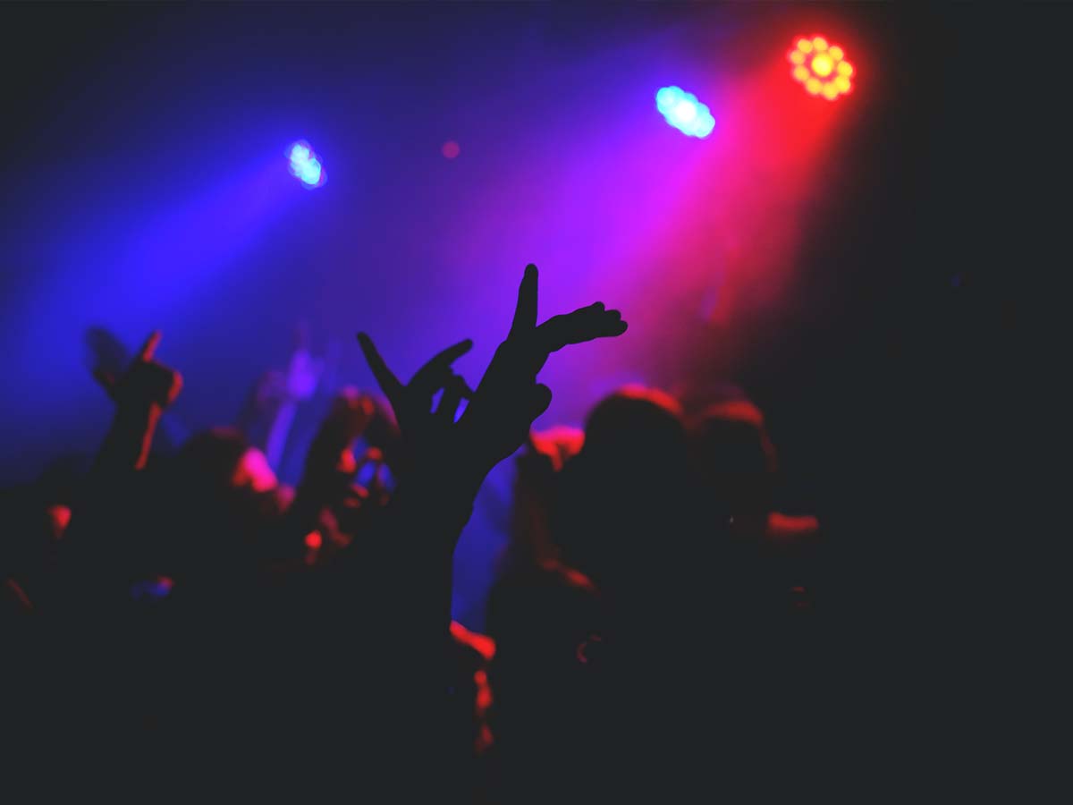 Hands in the air under a rig of purple lights in a club.