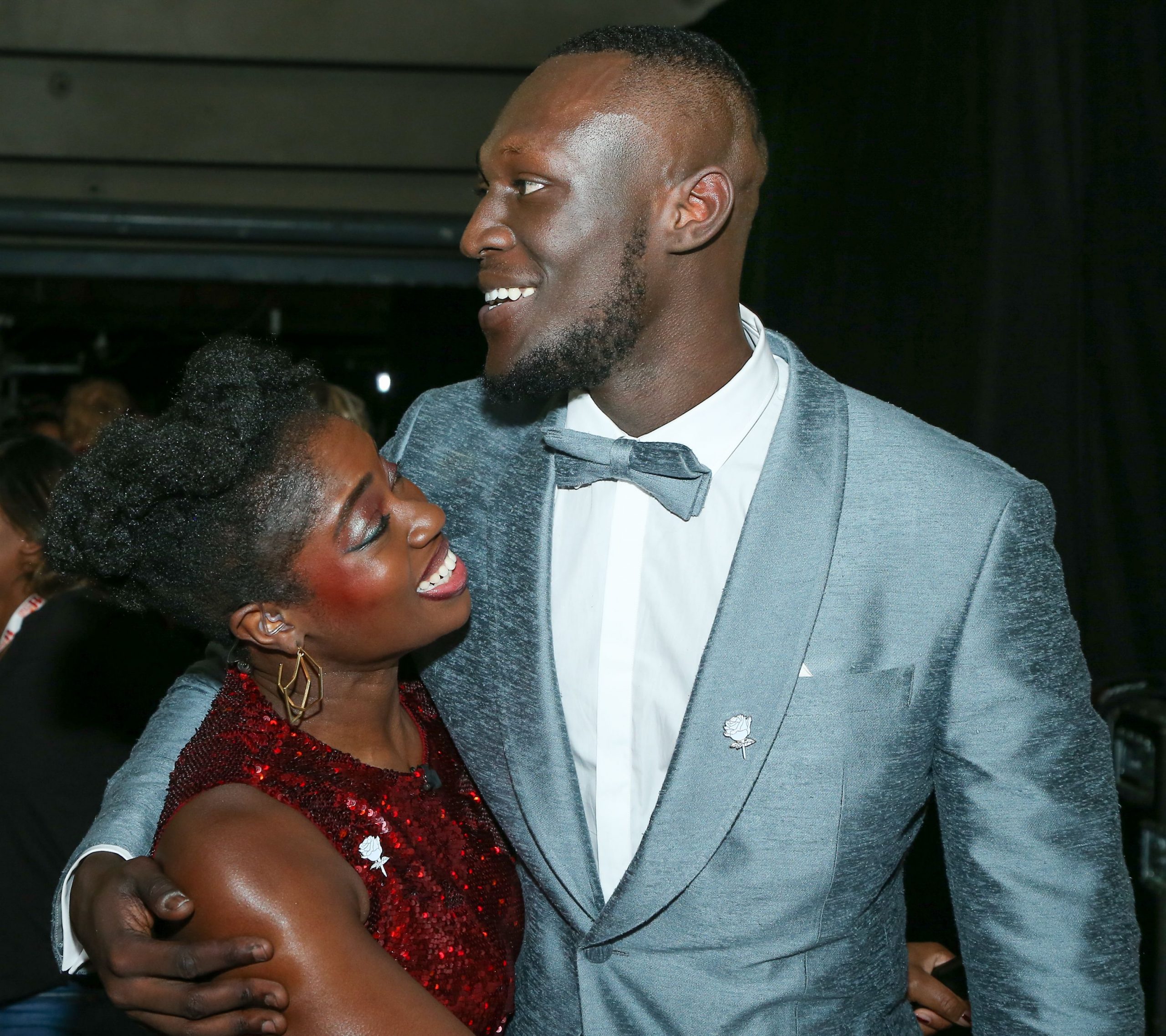 Clara Amfo in a red dress and Stormzy in a silver suit higging at a premier.