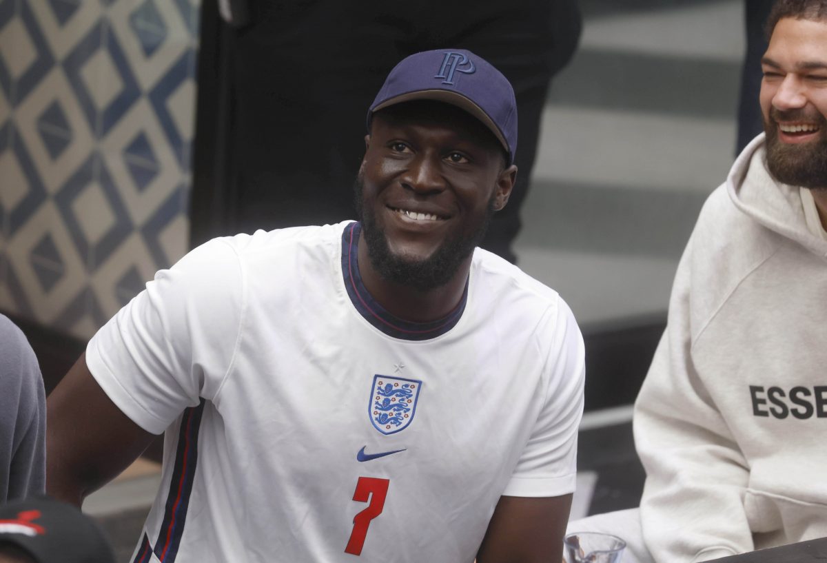 Sormzy at an England football game in 2021, he is dressed in a football T-shirt with the 3 lions logo and a red number 7 in the center. He also wears a blue baseball cap.