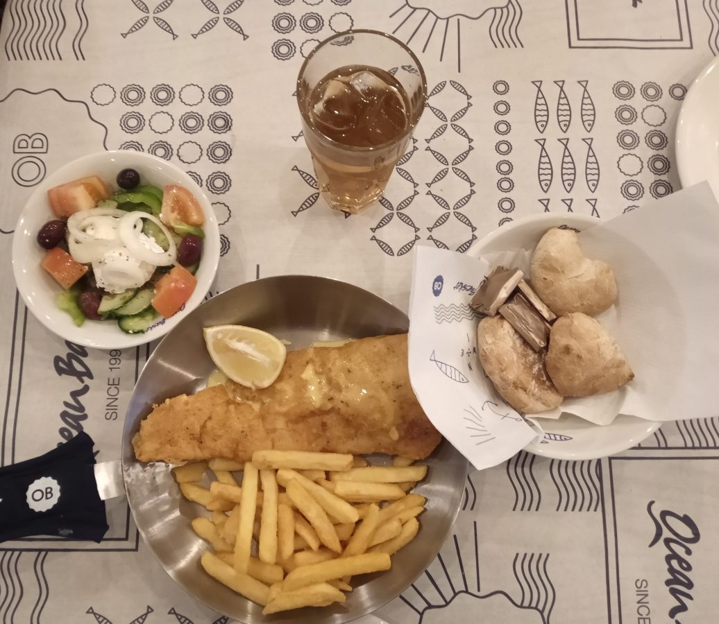 A plate of salad with chopped tomatoes, onions and cucumbers and black olives. There is a pan of battered fish and chips with a slice of lemon.  There is also a glass of ice tea with blocks of ice and a small bowl with bread and butter.