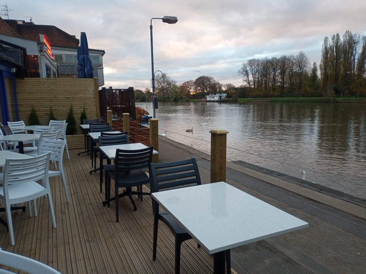 A picture of Kingston's riverside with swans. Next to riverside is the back of the restaurant with two and four seater tables and chairs for people to  sit and eat.
