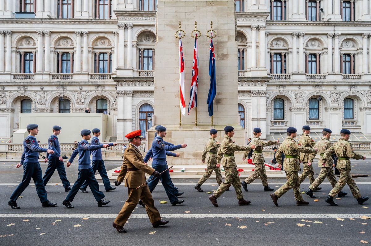 Cadets and armed forces wearing combat uniform march past the Cenotaph, where there are red poppy wreaths around it. There are also three British flags at the side of the Cenotaph.