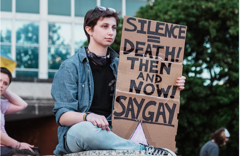 Person holding sign that says 'Silence = Death! Then and now! Say Gay'
