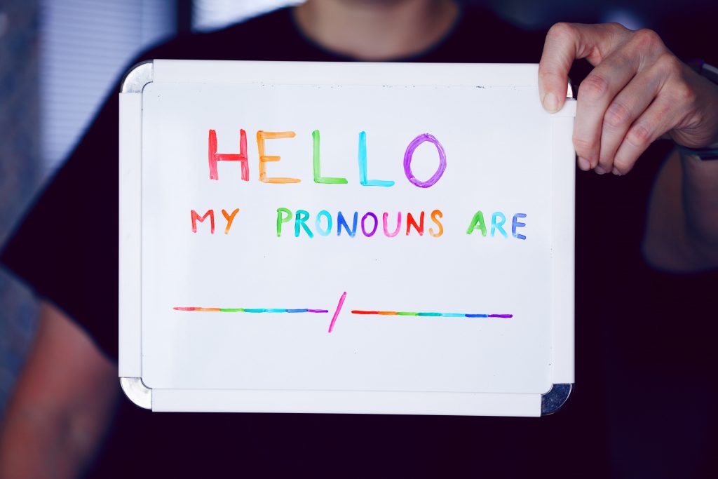 A person holding a small whiteboard with words: Hello my pronouns are, in coloured lettering representing the LGBT rainbow colours.